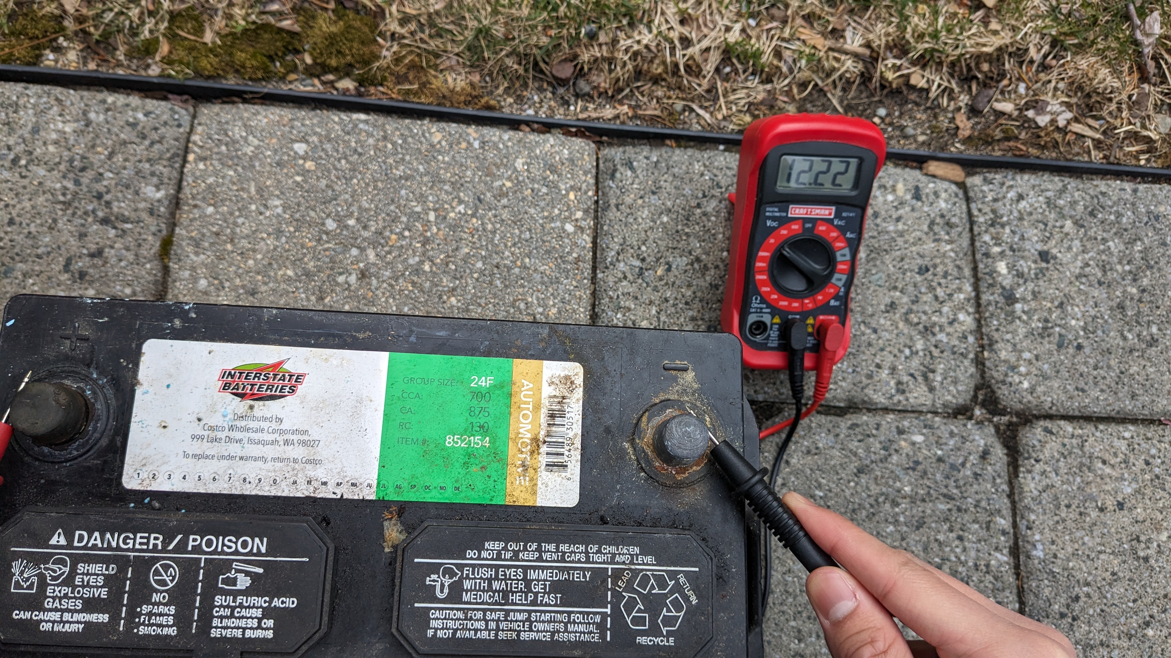 A lead-acid battery on the ground in front of a multimeter displaying a voltage reading of 12.32 V