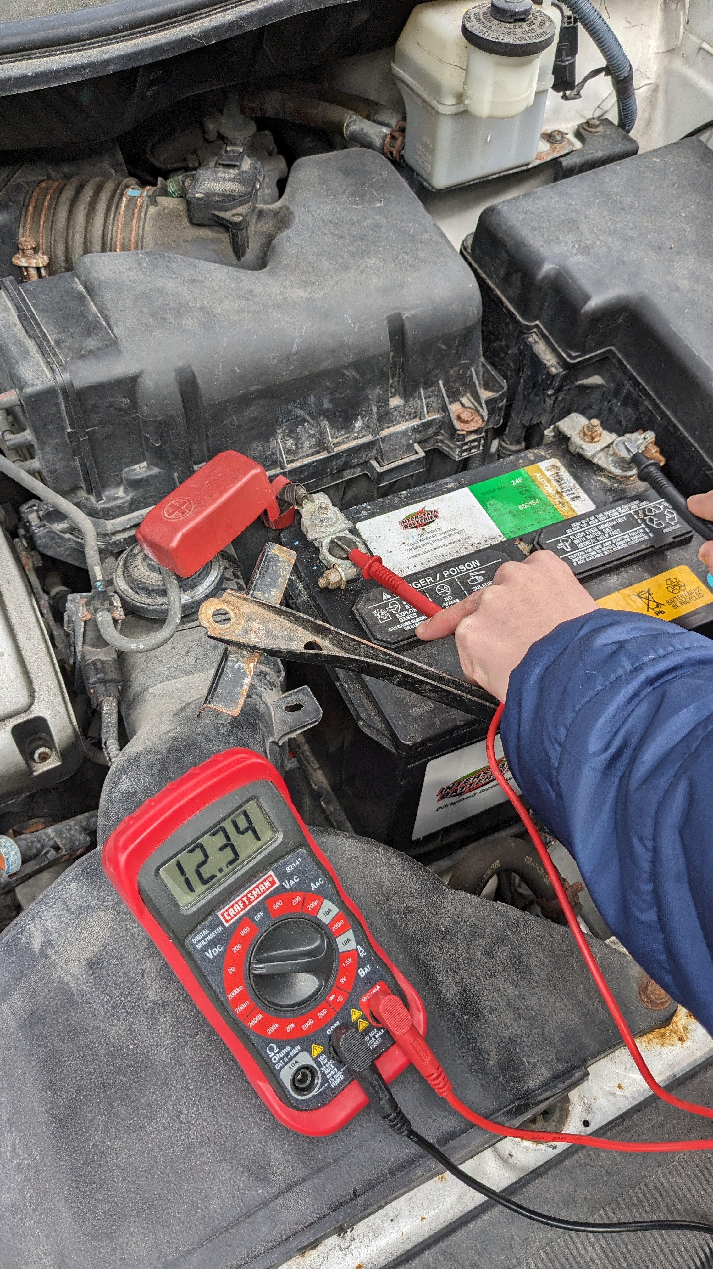 A lead-acid battery in a car with a multimeter in front displaying a voltage reading of 12.34 V